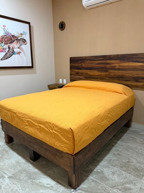 Una cama con estructura de madera y colcha amarilla en una habitación con cabecera de madera, aire acondicionado instalado en la pared y un cuadro enmarcado.