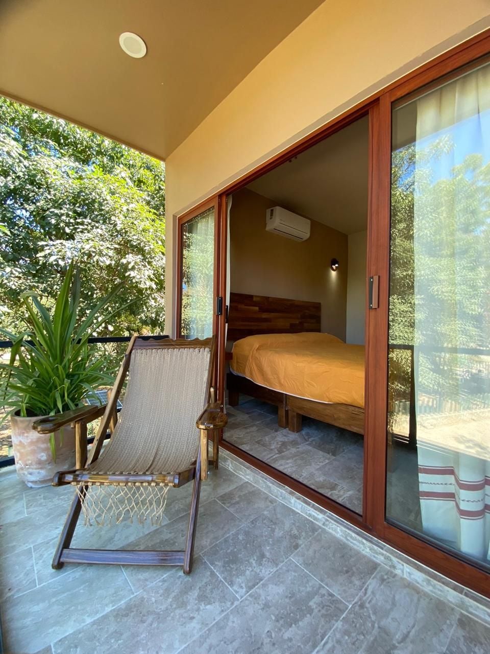 A room with a bed, a chair, and a sliding glass door leading to an outdoor area with a potted plant.