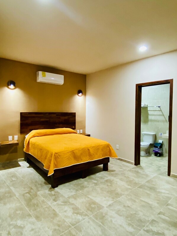 Un dormitorio con cama, aire acondicionado, mesita de noche y baño visible a través de una puerta abierta.