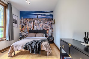 Un dormitorio con un mural de paisaje urbano, una cama con cabecera negra, una mesita de noche y un aparador con un adorno.