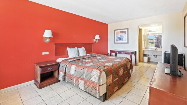 A hotel room with a red accent wall, a bed with a quilt, bedside lamps, a desk with a computer, and a bathroom visible through an open door. - Luxury Inn By Hotel O Hattiesburg, MS (Hattiesburg)