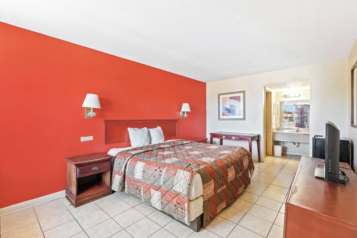 Une chambre d’hôtel avec un mur d’accent rouge, un lit recouvert d’une courtepointe, des lampes de chevet, un bureau avec un ordinateur et une salle de bain visible à travers une porte ouverte.