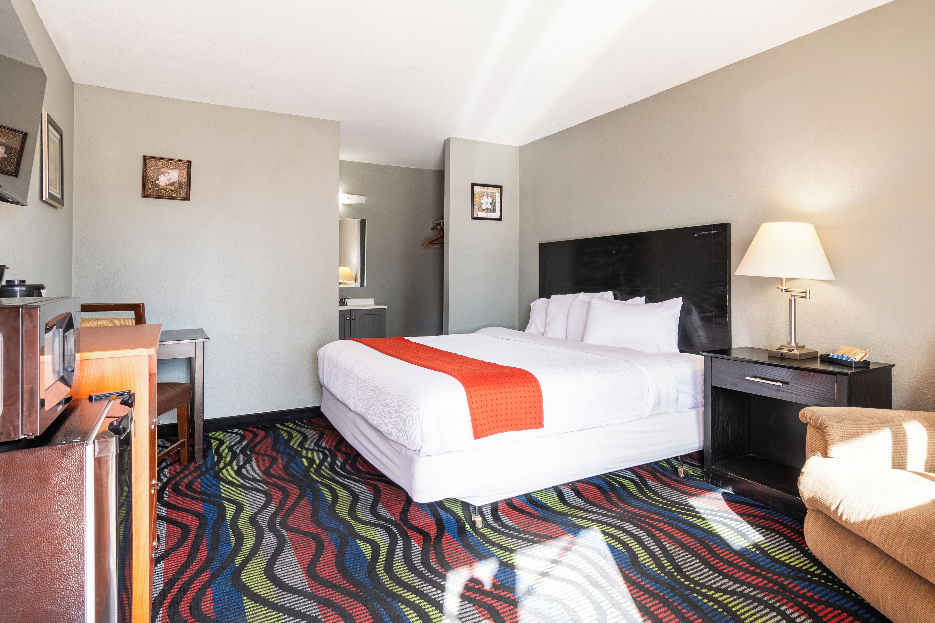 Una habitación de hotel con cama, escritorio, silla, mesita de noche, lámpara y espejo.