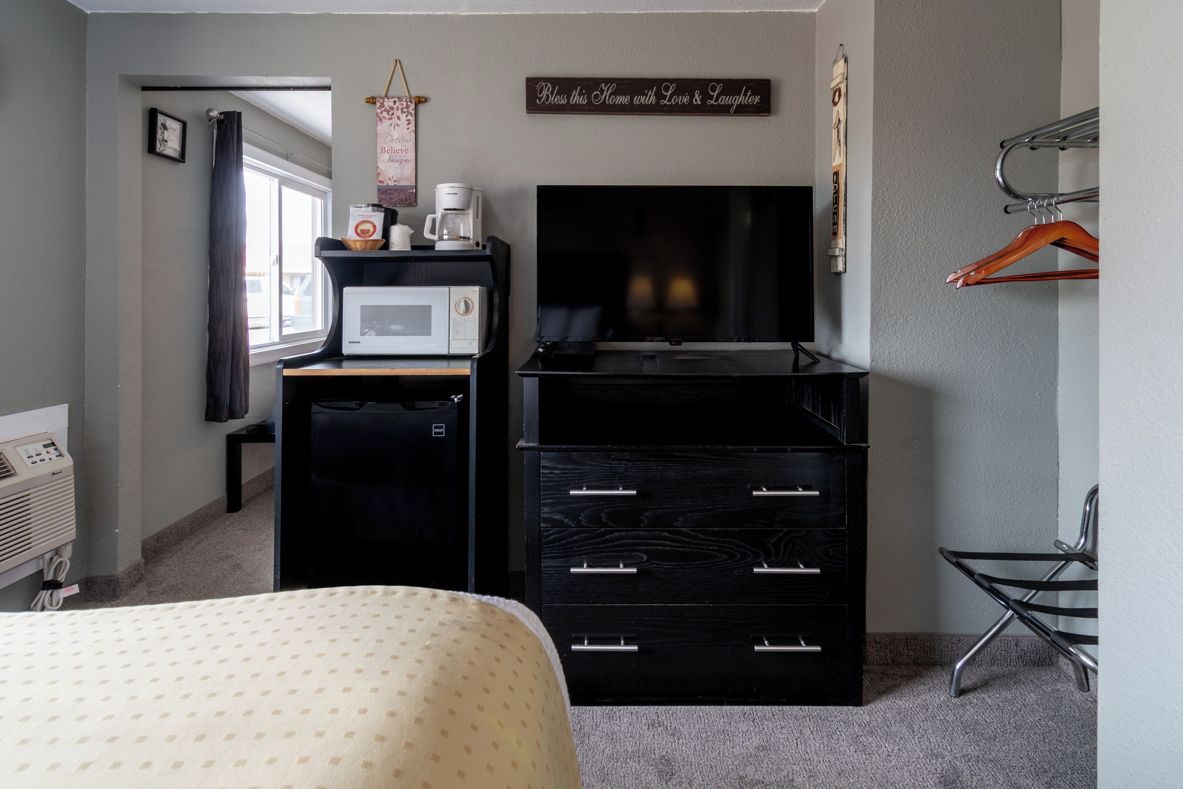 Habitación de hotel con cama, escritorio con microondas, televisor, cómoda y perchero.