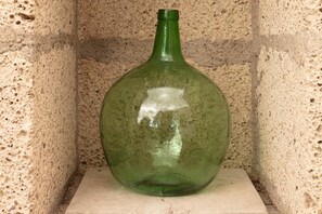 Une bouteille en verre vert au goulot étroit, posée sur une corniche en pierre, adossée à un mur à la texture marquée.