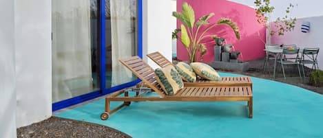 A sun lounger with cushions, a small table with chairs, and a pink wall.