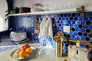 Une cuisine avec un revêtement mural en carreaux hexagonaux bleus, une étagère blanche où sont disposés des verres, un panier de fruits et un porte-ustensiles doré.