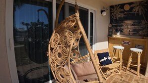 Une chaise suspendue en osier avec un coussin, un fauteuil à bascule en osier et un tabouret de bar dans une pièce dotée d’une porte en verre et d’une fresque murale représentant une plage.