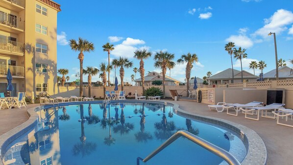 A swimming pool with lounge chairs and palm trees, surrounded by buildings. - Beachside 1st Floor Condo with Patio,Heated Pool & Gas BBQ – 3 Min Walk to Beach (Daytona Beach Shores)