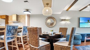 Un coin repas avec des chaises en osier, une table ronde agrémentée d’un centre de table et un téléviseur diffusant une scène de plage.