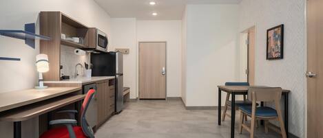A compact kitchenette with a microwave, sink, and wooden cabinets. There is a desk with a chair, a dining table with chairs, and a patterned rug.