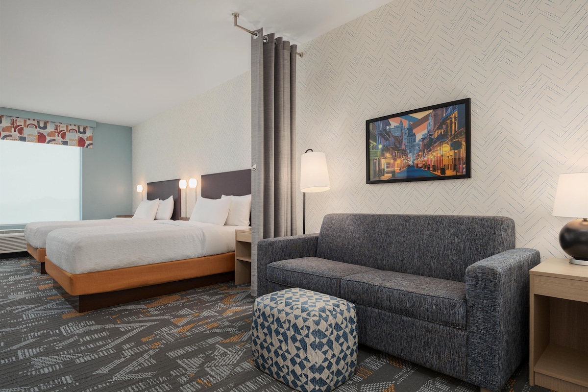 A hotel room with a bed, a sofa, a patterned ottoman, a nightstand, a lamp, and a cityscape painting on the wall.