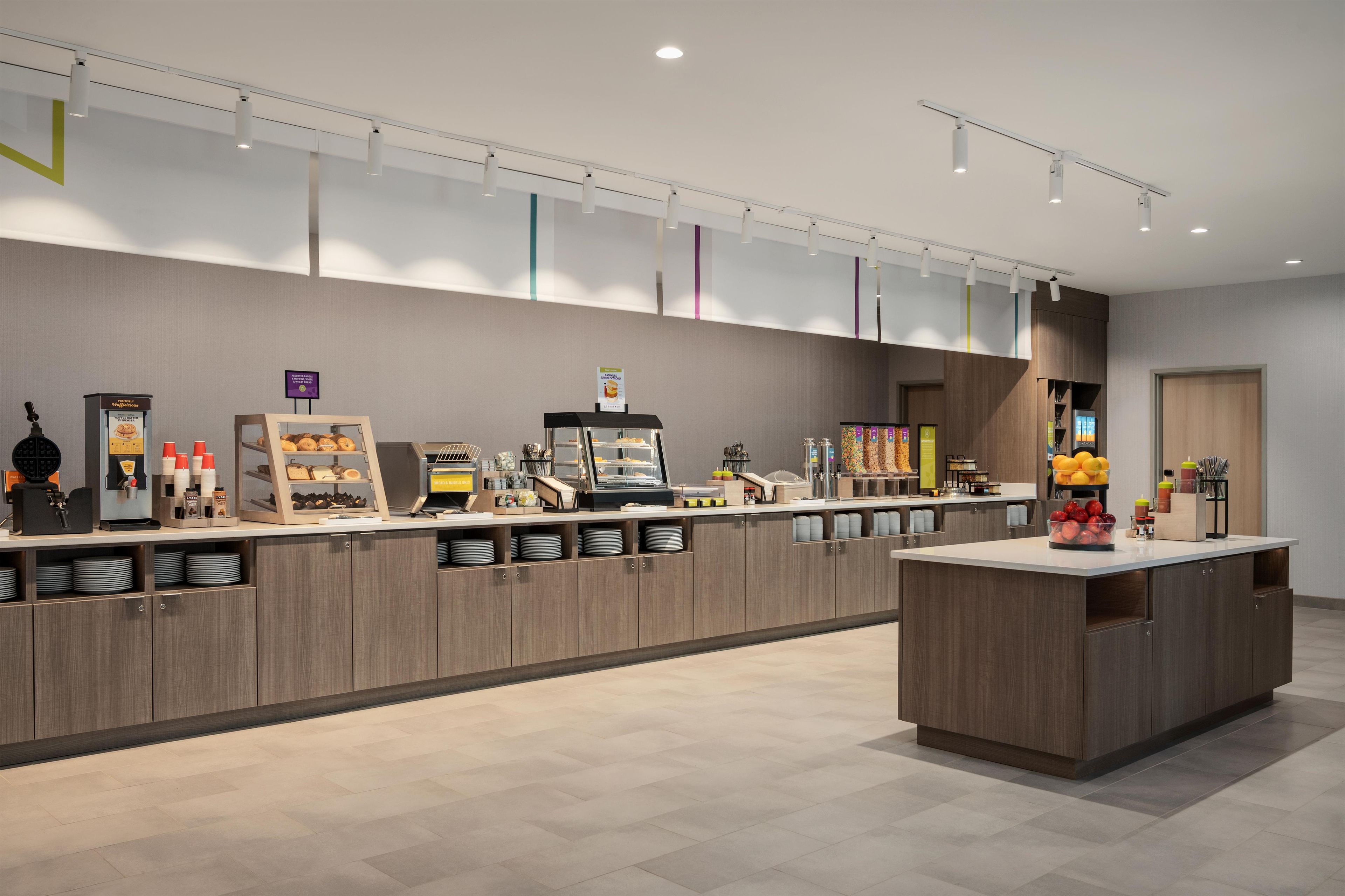 Un área de recepción moderna de hotel con mostrador, máquinas de café y una exhibición de frutas.