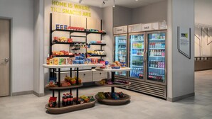 A snack display with shelves and a table, a refrigerator with beverages, and a sign stating 'HOME IS WHERE THE SNACKS ARE'. - Home2 Suites By Hilton Lafayette I-10 (Lafayette)