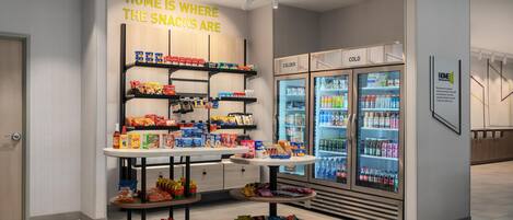 A snack display with shelves and a table, a refrigerator with beverages, and a sign stating 'HOME IS WHERE THE SNACKS ARE'.