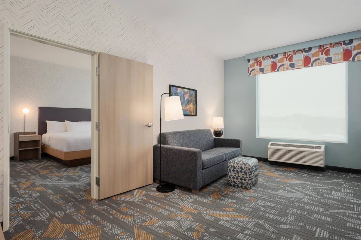 A hotel room with a bed, a sofa, a small table, a lamp, a picture on the wall, and a large window.