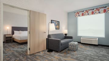 A hotel room with a bed, a seating area with a chair and ottoman, a standing lamp, a wall-mounted picture, and a large window.