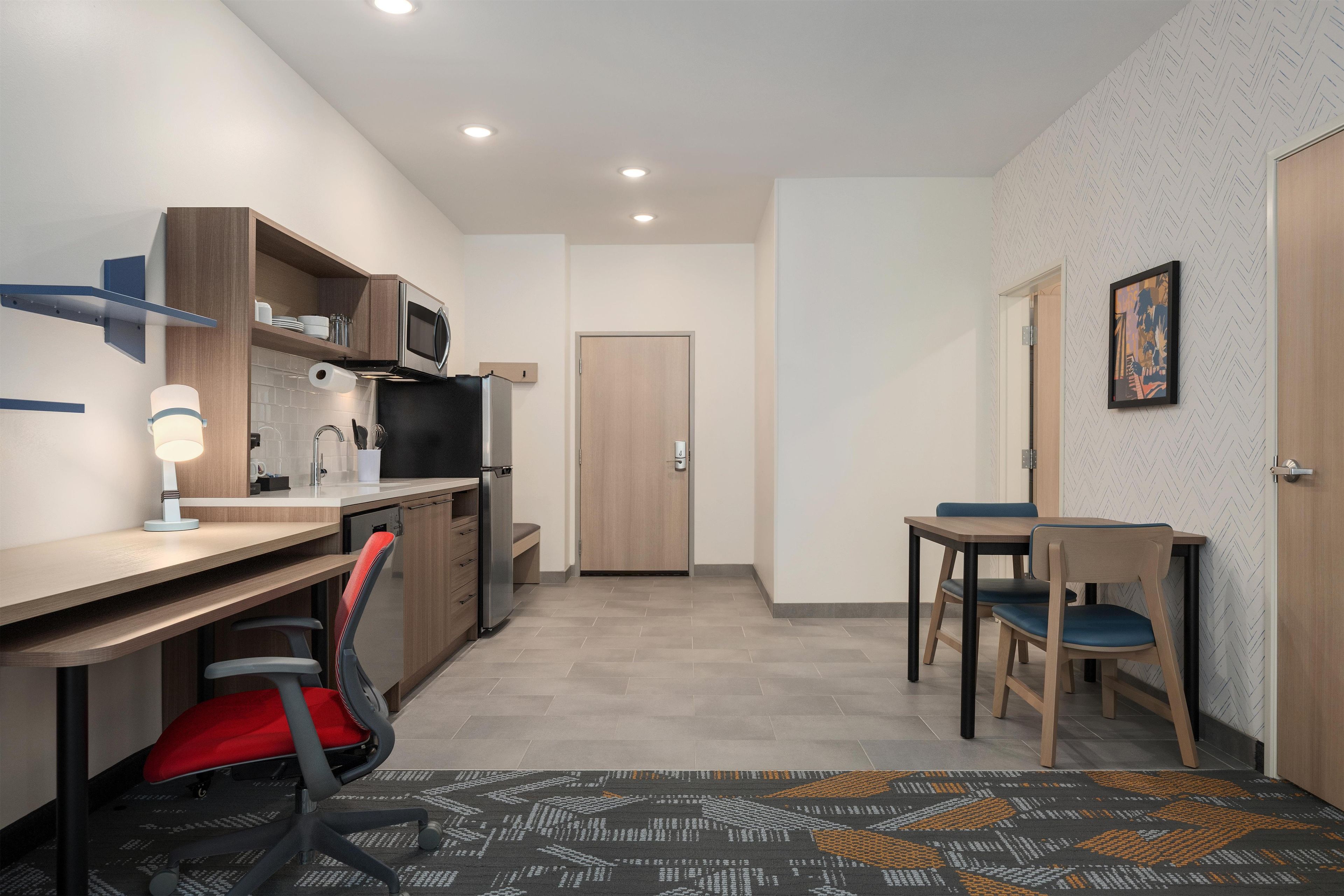 Una kitchenette compacta con microondas, pileta y muebles de madera. Hay un escritorio con silla, una mesa de comedor con sillas y un alfombra estampado.