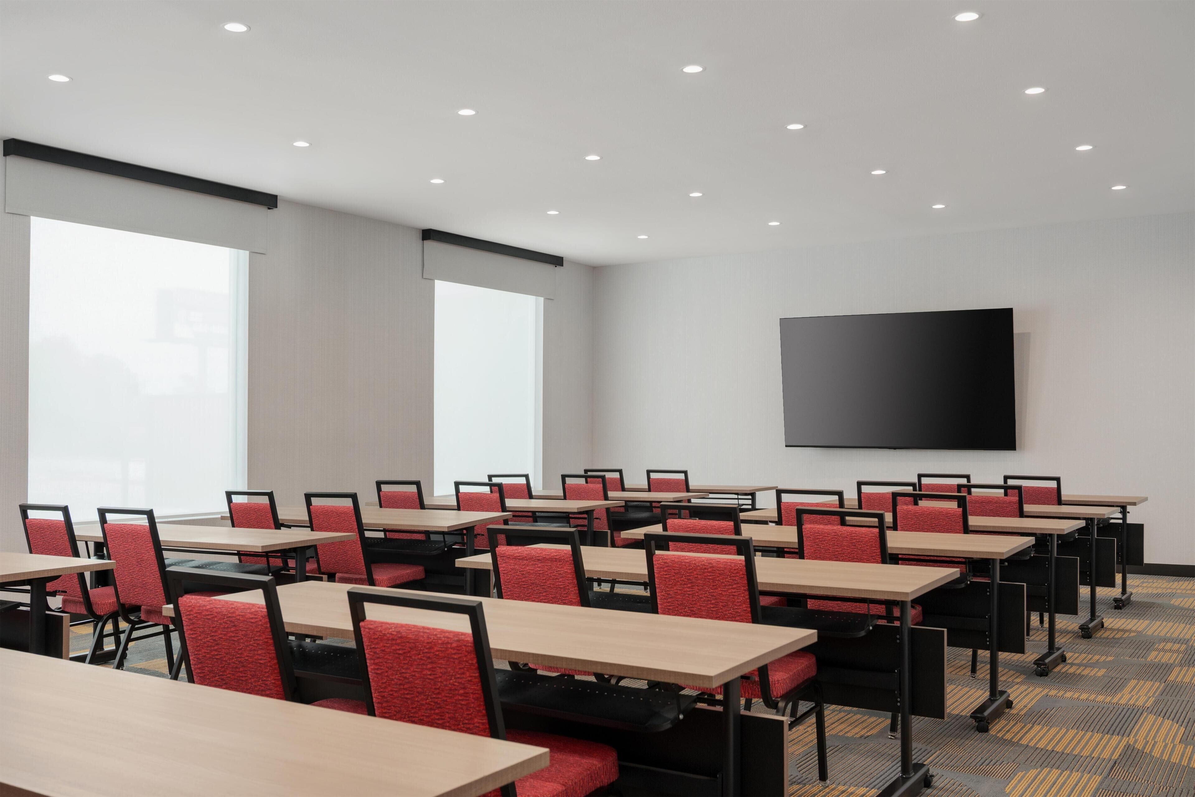 Un salón de conferencias con varias mesas y sillas, un televisor de pantalla plana grande y ventanas que dejan entrar luz natural.
