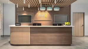 A modern hotel reception desk with a wooden finish, two monitors, and a sign that reads 'HOME2'. - Home2 Suites By Hilton Lafayette I-10 (Lafayette)