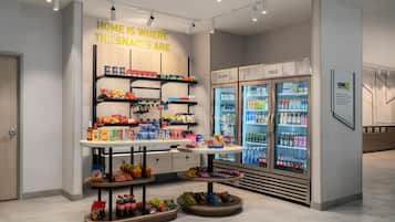 Una exposición de bocadillos con estantes y una mesa, un refrigerador con bebidas, y un cartel que dice 'HOME IS WHERE THE SNACKS ARE'.