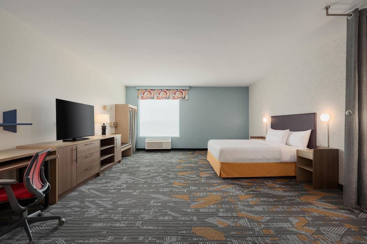A hotel room with a bed, desk, TV, and a window with curtains.