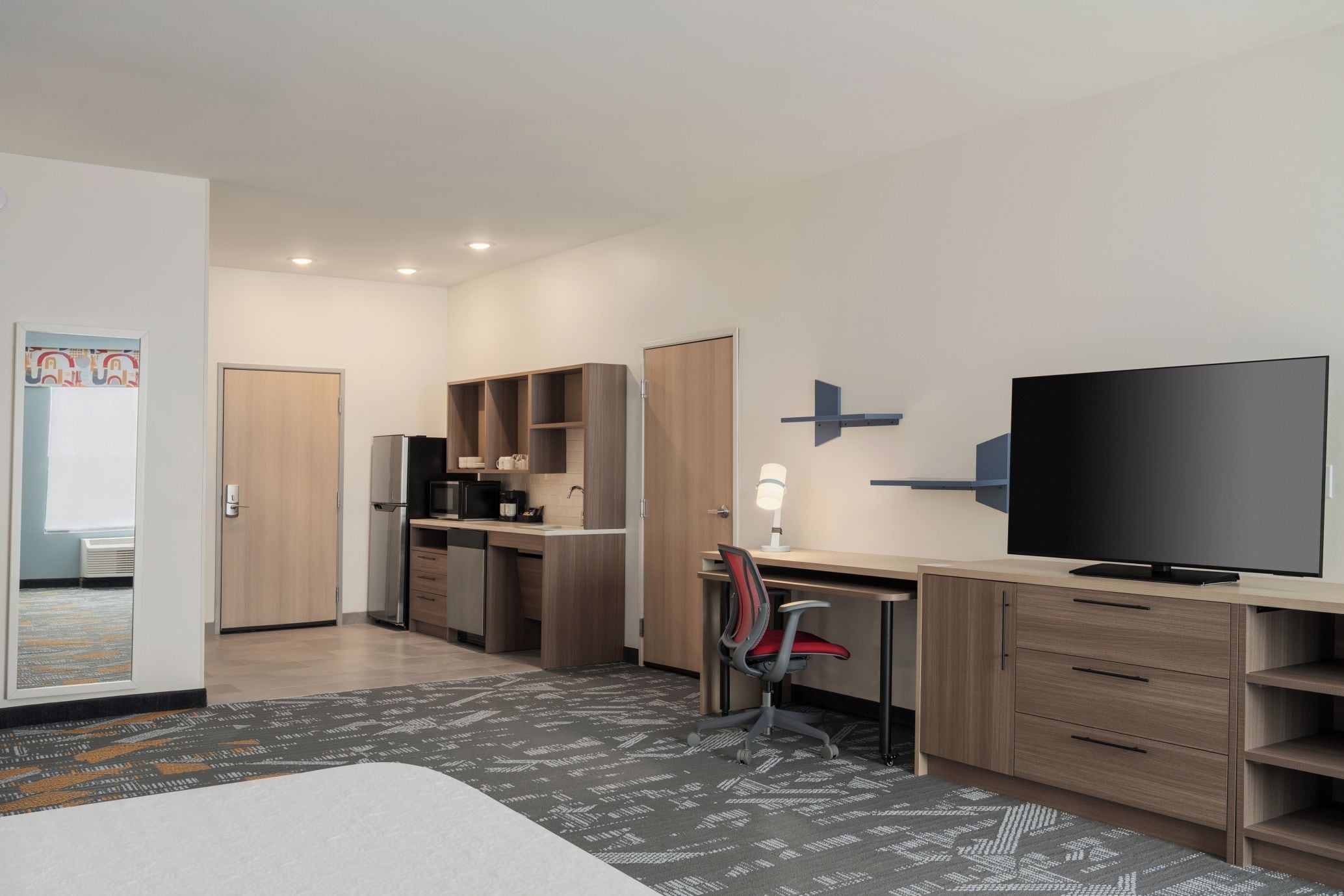A modern hotel room with a kitchenette, a flat-screen TV, a desk with a chair, and a patterned rug.