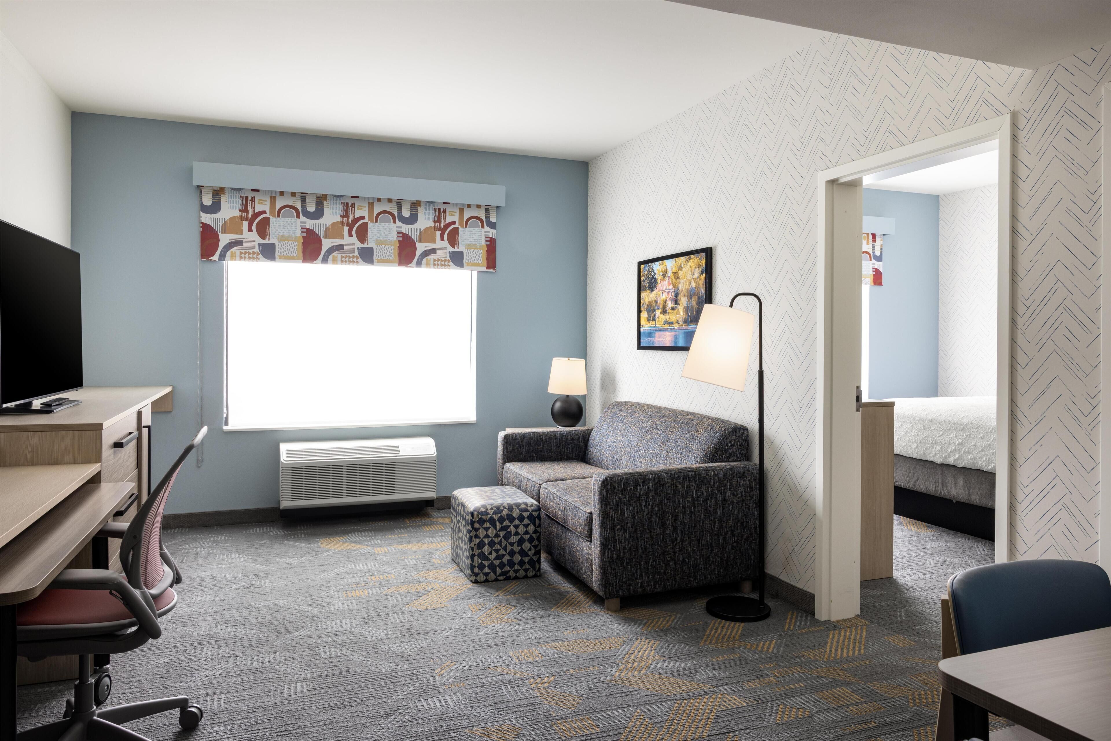 A hotel room with a desk, chair, sofa, ottoman, lamp, and a bed.