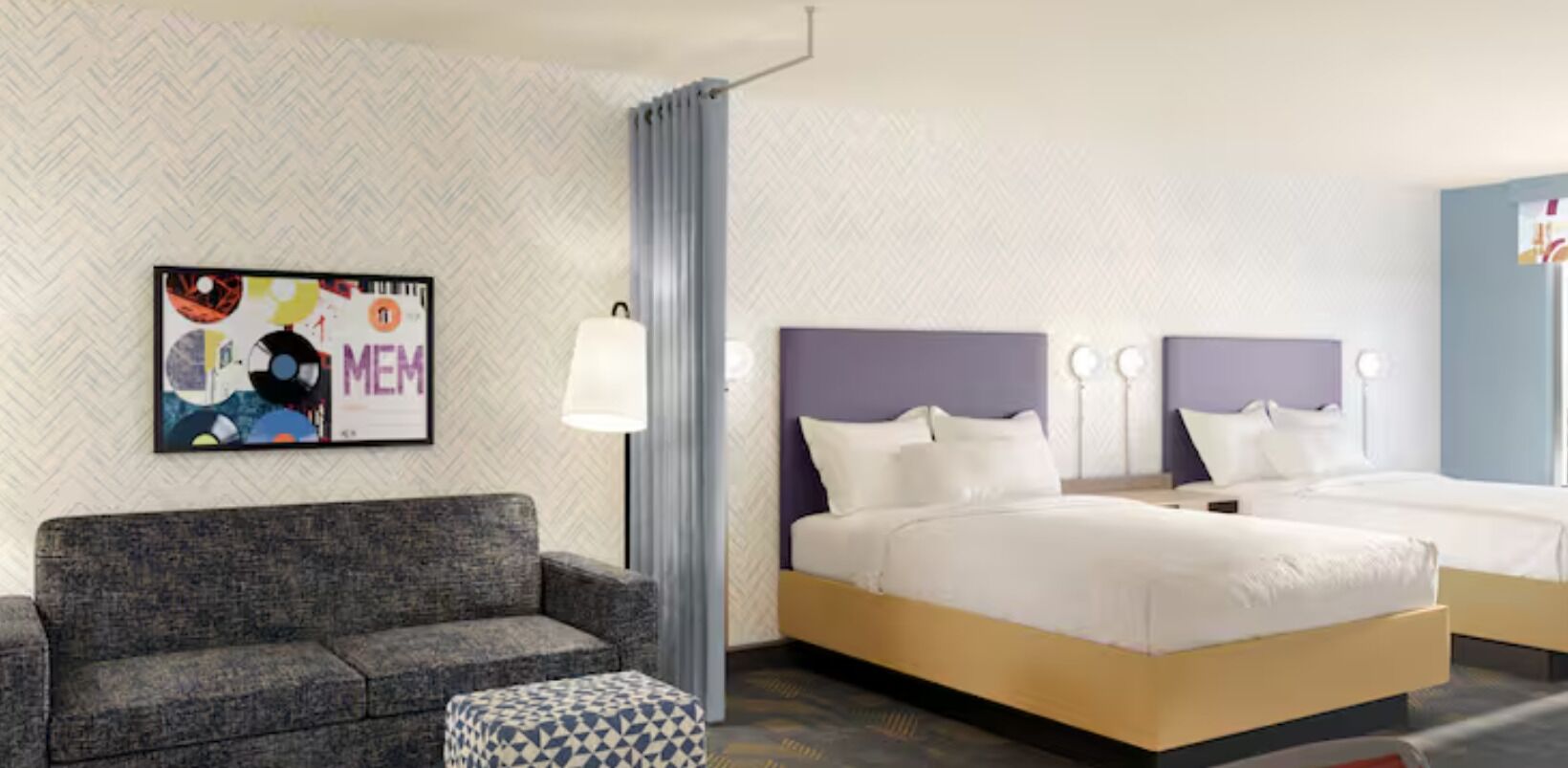 Hotellihuone, jossa on sohva, sänky, yöpöytä, lamppu ja kuviollinen tapetti yhdellä seinällä.