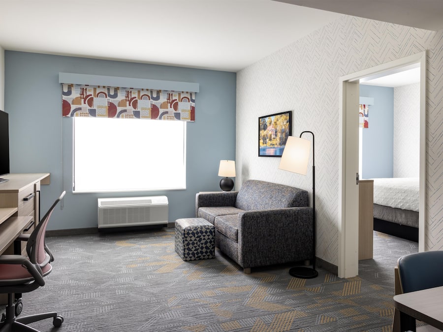 A hotel room with a desk, chair, sofa, ottoman, lamp, and a bed.