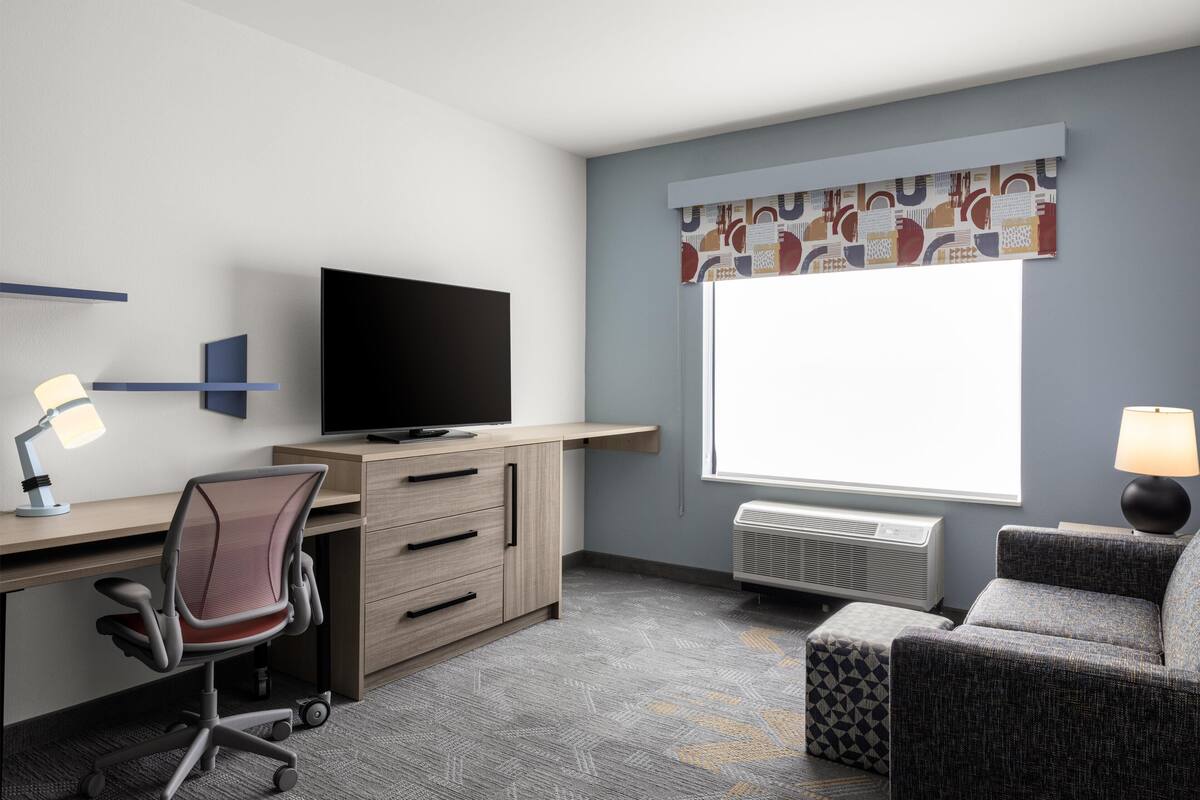 Habitación de hotel con escritorio, silla, televisor, ventana con cortinas y un sofá.