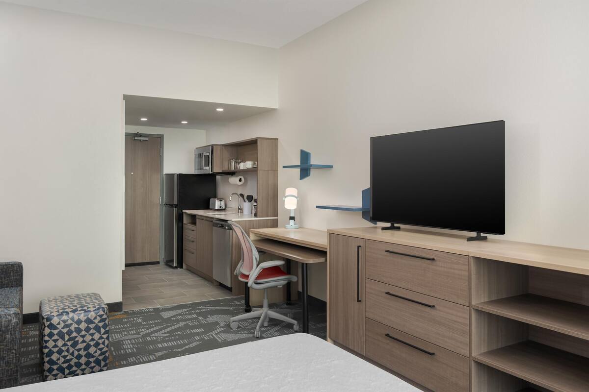 A modern hotel room with a bed, a desk, a chair, a TV, a small kitchenette, and a bathroom.