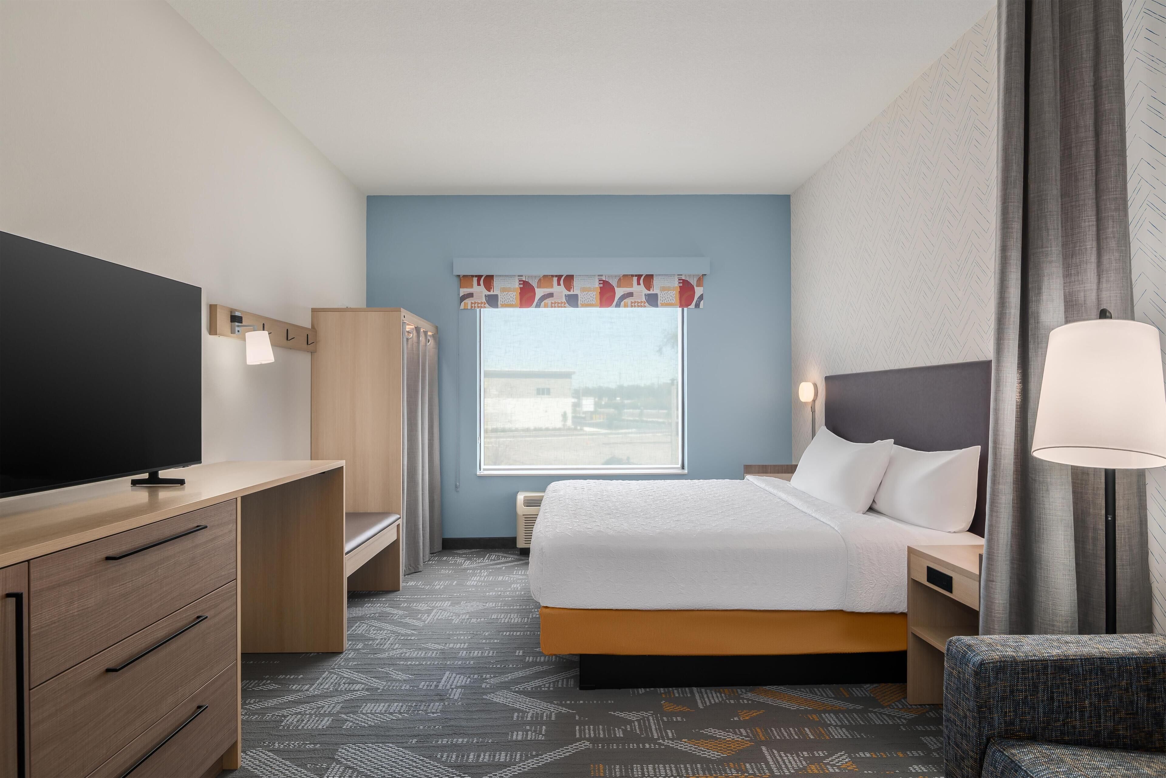 Habitación de hotel con cama, mesita de noche, lámpara, televisor, armario y ventana con vistas.