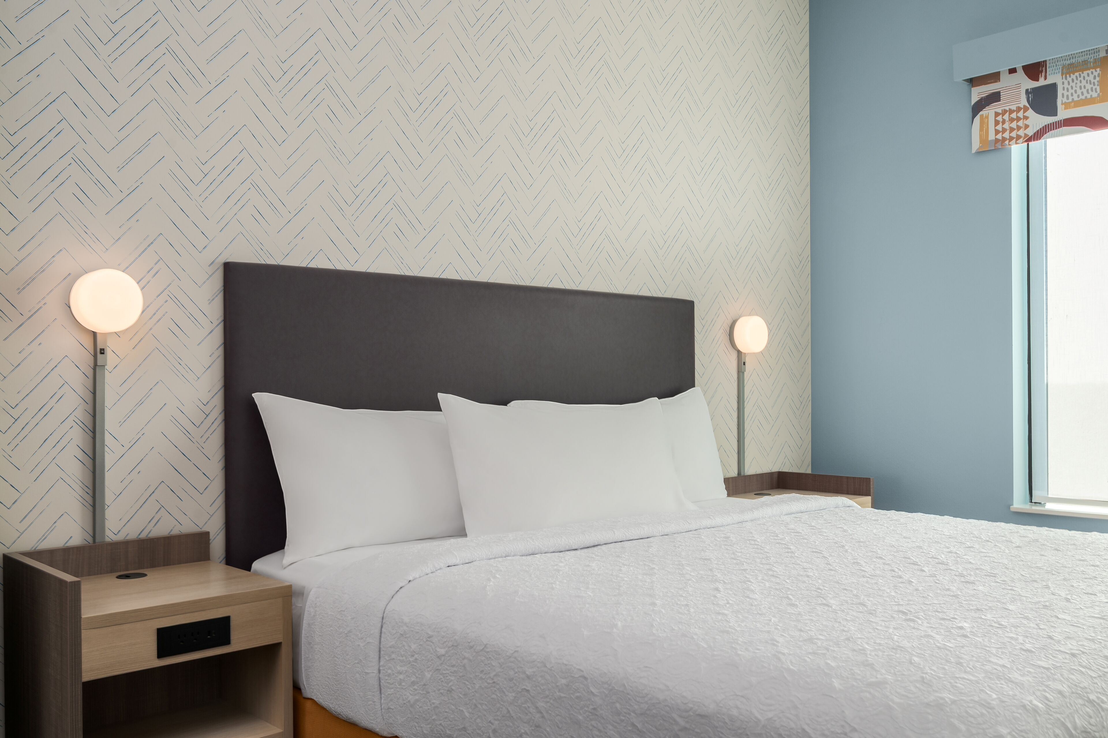 A neatly made bed with white linens, two bedside tables with lamps, and a herringbone-patterned wall.