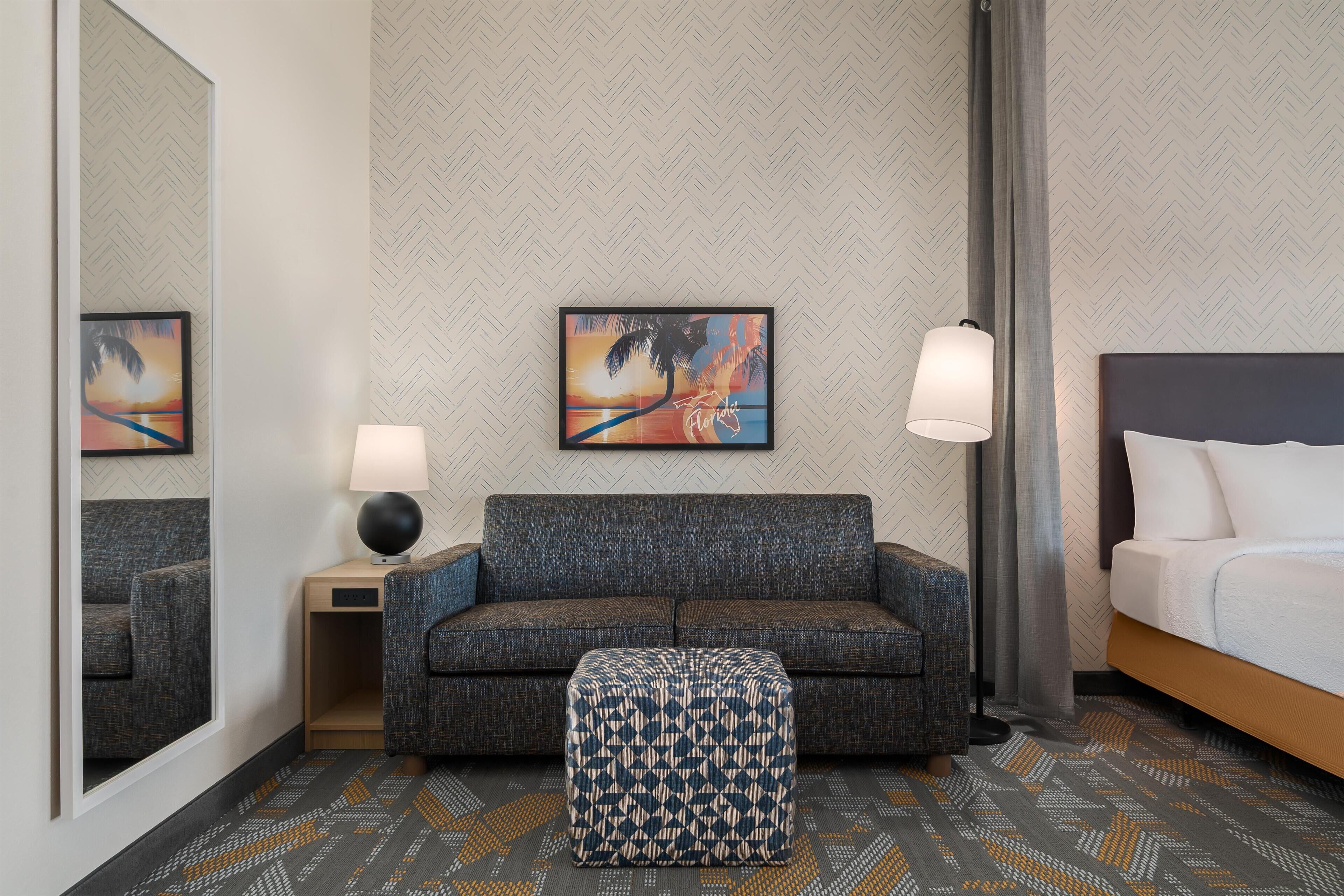 A hotel room with a sofa, ottoman, two lamps, a bed, a mirror, and two wall art pieces.