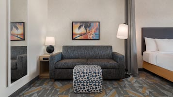 A hotel room with a sofa, ottoman, two lamps, a bed, a mirror, and two wall art pieces.