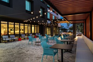 An outdoor seating area with tables and chairs, a covered section with a wooden ceiling, and a building with large windows in the background. - Home2 Suites By Hilton Riverview Tampa (Riverview)