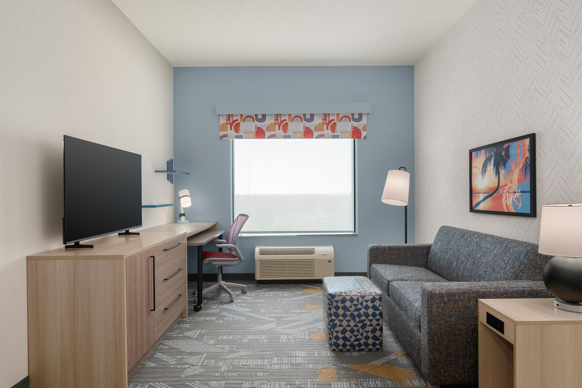 Una habitación de hotel moderna con televisión de pantalla plana, un escritorio, un sofá cómodo, una alfombra estampada y un ventanal con vistas.