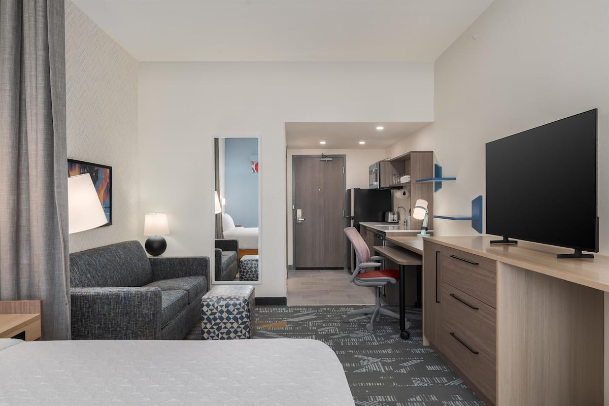 A hotel room with a bed, a sofa, a desk with a chair, a television, and a kitchenette.