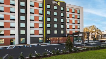 Moderni hotellirakennus, jossa on selkeästi näkyvä «HOME 2» -kyltti, katettu sisäänkäynti ja pysäköintialue, jossa on myös invapaikkoja.
