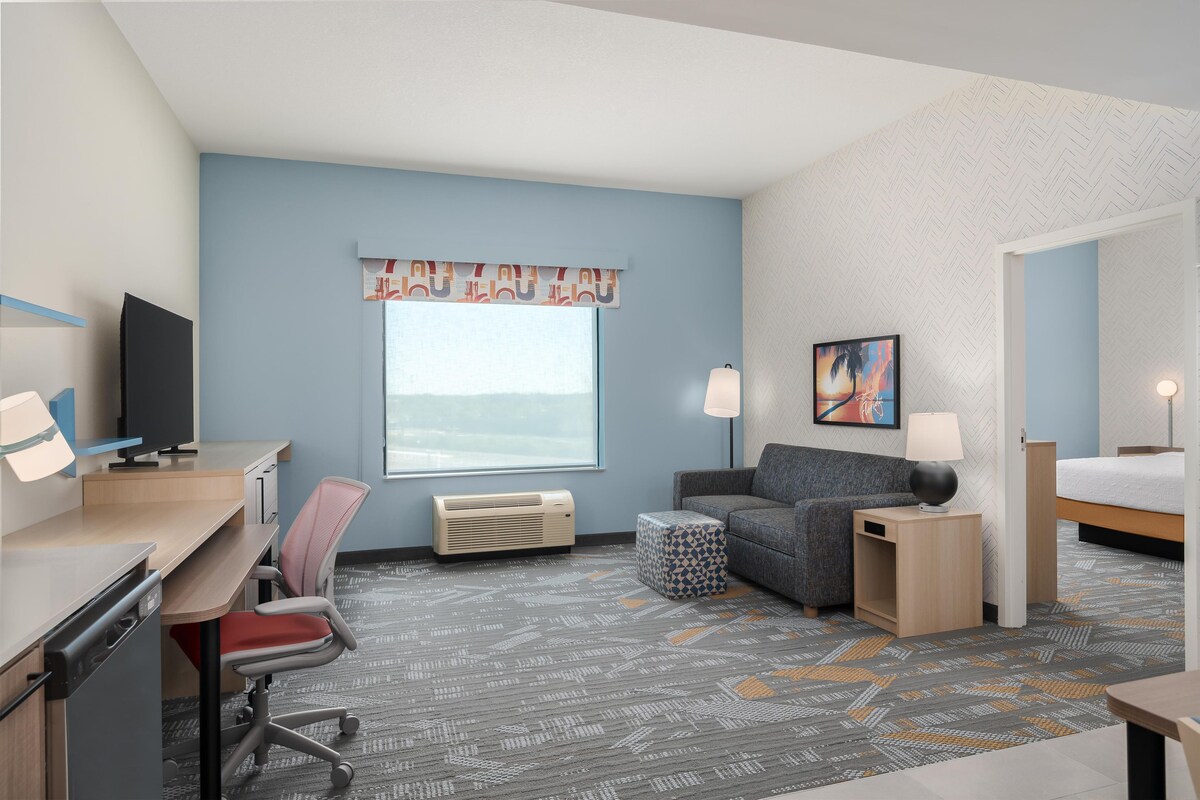 Habitación de hotel con escritorio, silla, sofá, mesita de noche, lámpara y cama.