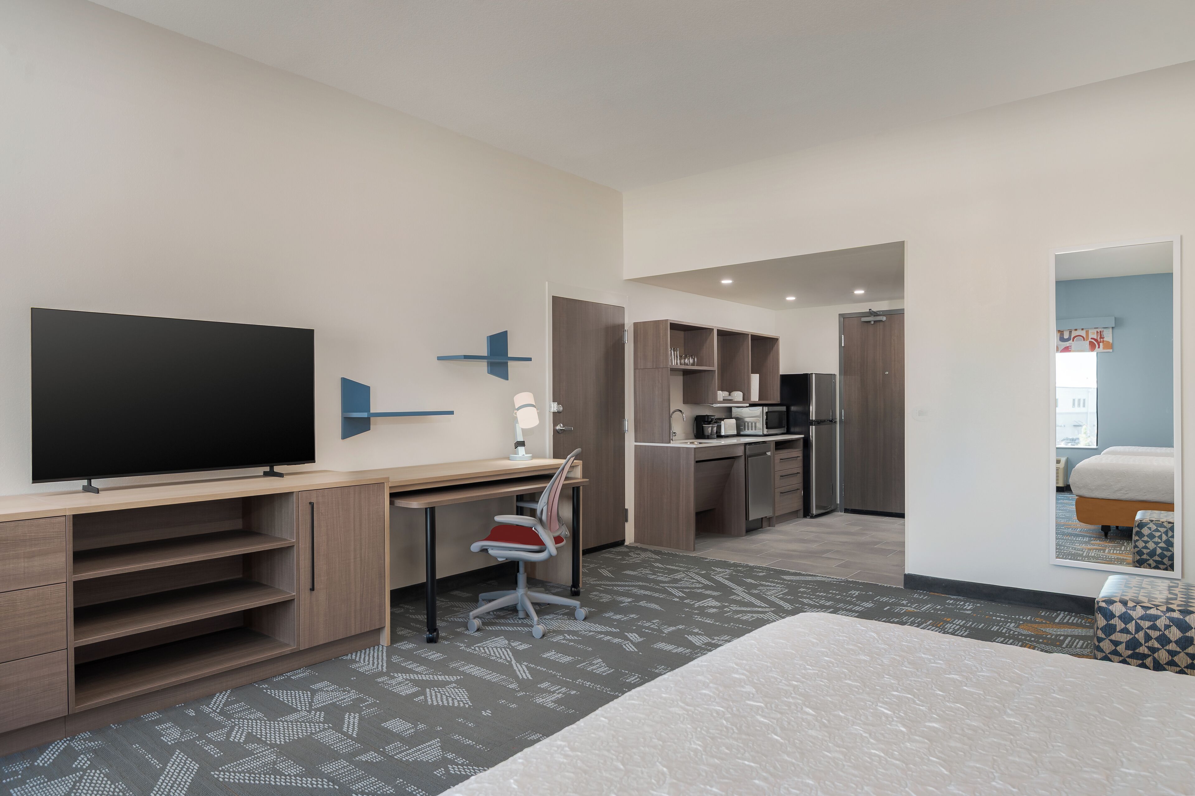 A modern hotel room with a large flat-screen TV, a desk with a chair, a kitchenette, and a bed with a patterned rug.