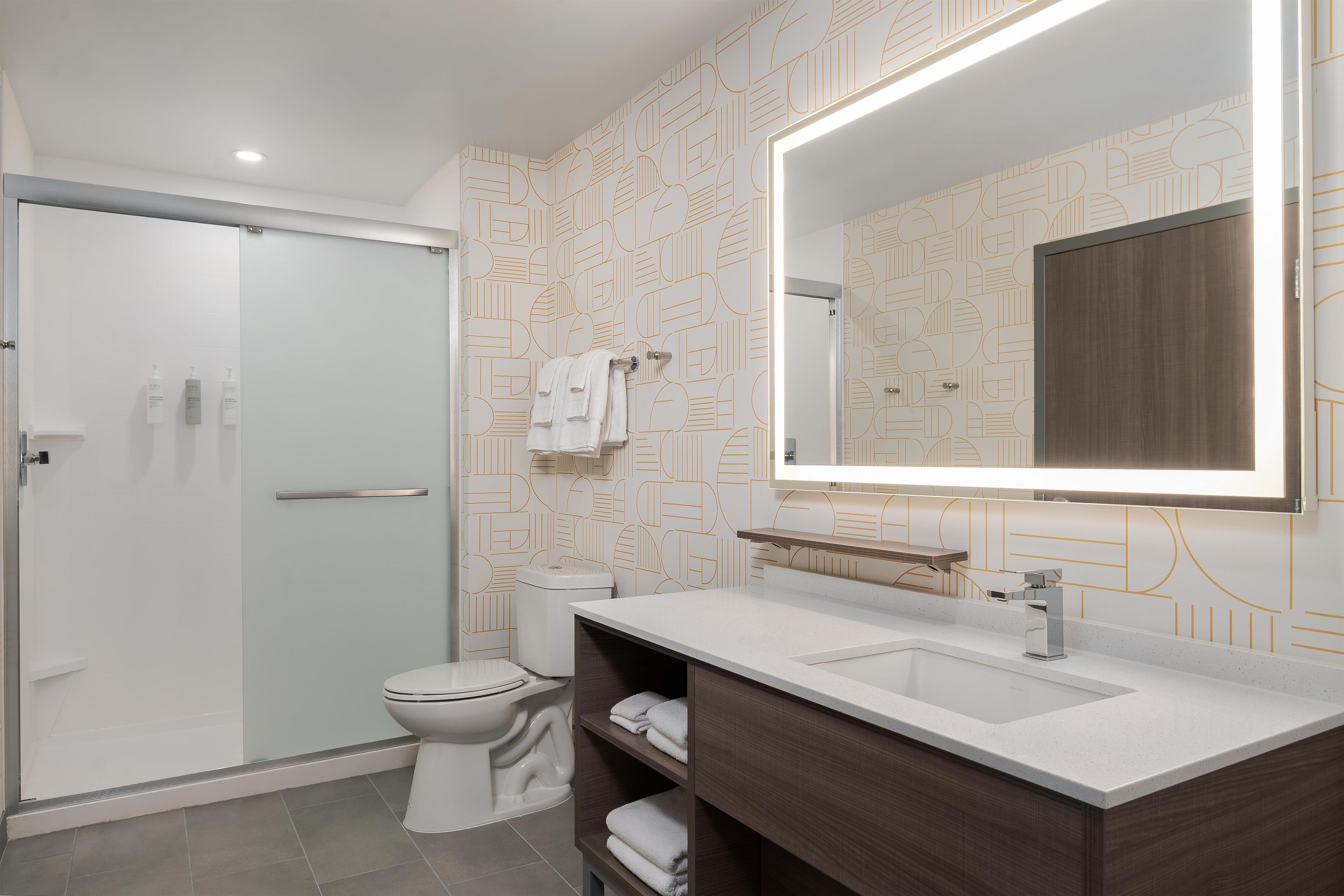 A modern bathroom with a glass shower enclosure, a toilet, a sink with a mirror, and patterned wallpaper.