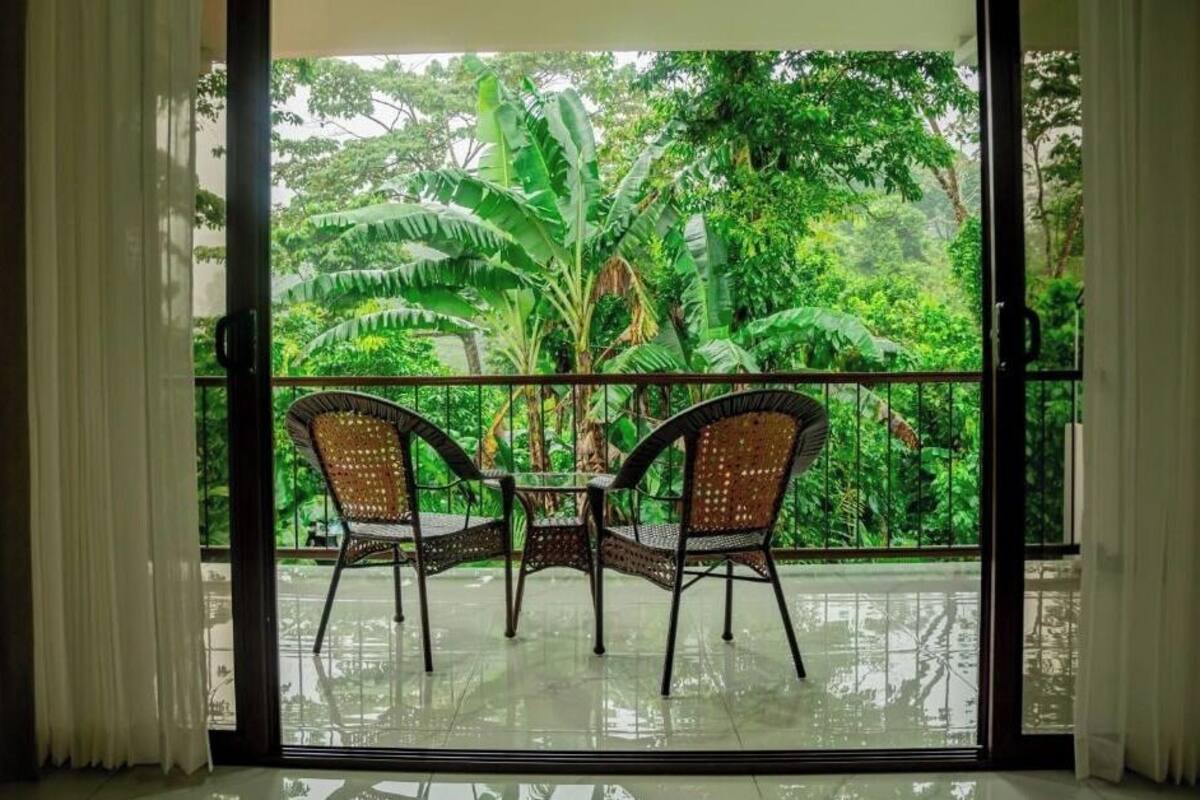 Varanda com duas cadeiras de vime e uma mesa, com vista para vegetação exuberante e bananeiras.