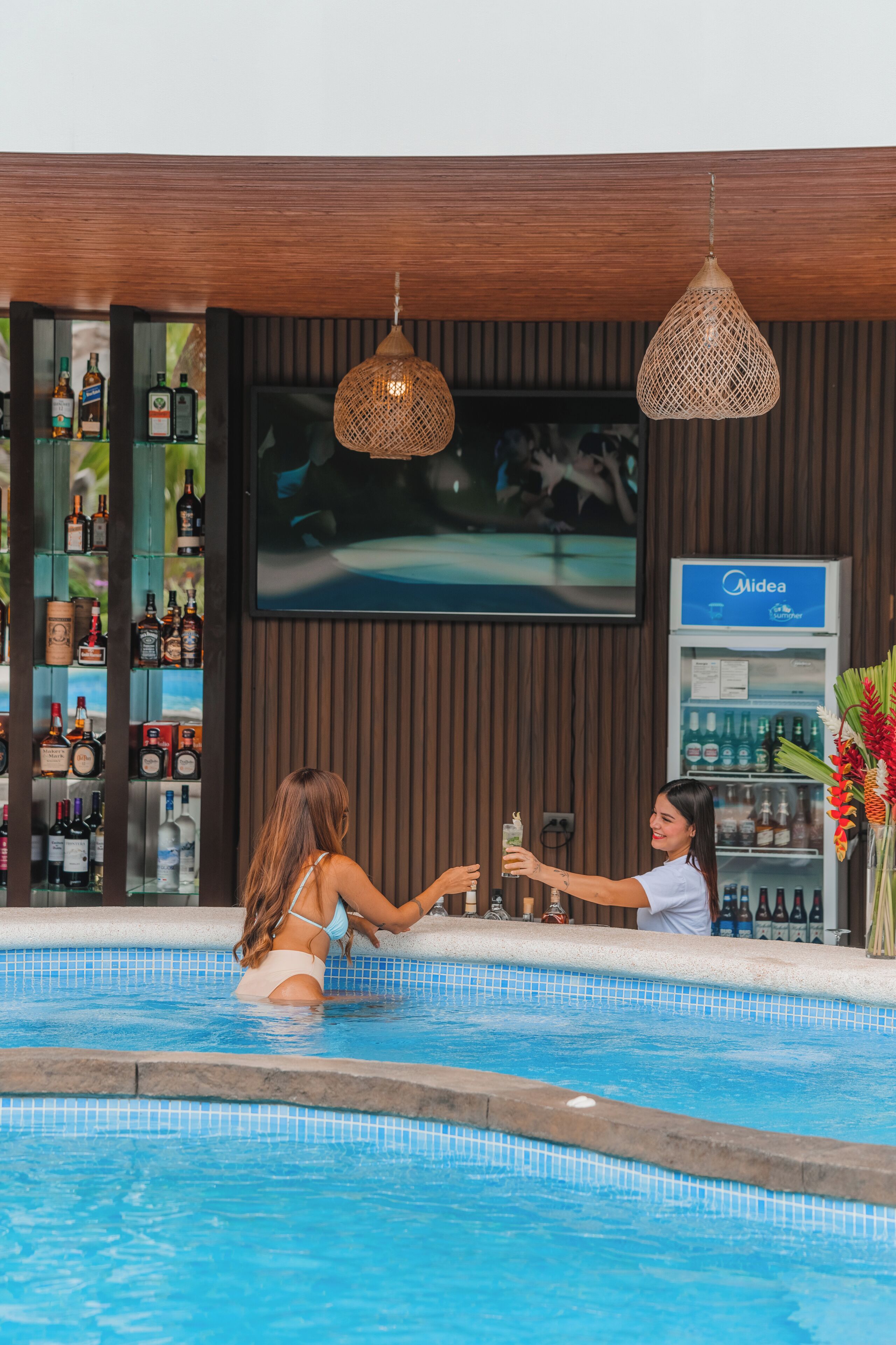 A poolside bar area with a television, bar shelves stocked with bottles, and a minibar.