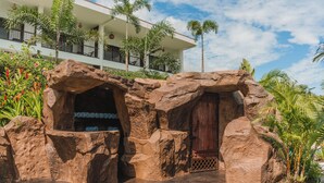 A rock formation entrance with a wooden door, surrounded by tropical plants and palm trees, leading to a modern building with balconies. - Hotel Sanctuarium Adults Only (La Fortuna)
