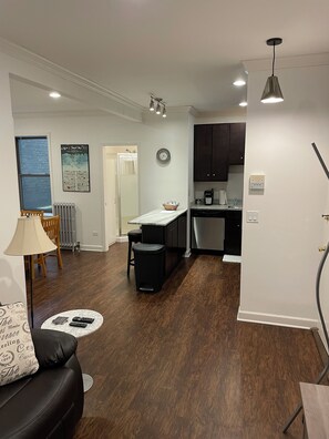 A modern kitchen with dark cabinets, a white countertop, and a black trash bin. There is a dining area with a black leather couch and a small round table. - Updated 1 bedroom in the best location! (Chicago)