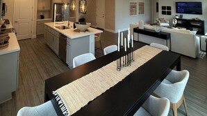 A modern kitchen with a central island, dining area with a black table and white chairs, and a living room with a sofa and TV.