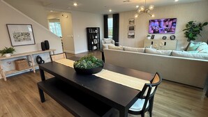 A modern living room with a dining area, featuring a wooden floor, a black dining table with a woven placemat, a sofa, and a television mounted on the wall.