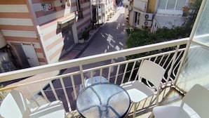 A balcony with a glass table and four white chairs, overlooking a street with buildings and parked cars. - Comfort Apartments Igalo 60 m From the Sea (Igalo)